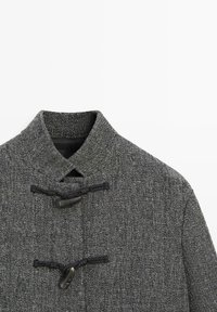 Cappotto in tessuto grigio con colletto alto, chiusure a fibbia e superficie testurizzata. Il design presenta un motivo sottile su tutta la superficie.