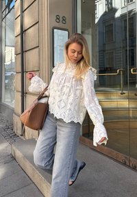 Weiße bestickte Bluse mit Rüschkragen und -ärmeln, kombiniert mit hellgrauen Jeans. Braune Tasche über der Schulter, stehend vor einem gläsernen Eingang.