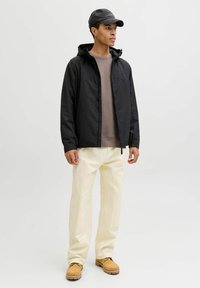 Veste légère noire avec capuche, portée sur un sweat-shirt taupe, associée à un pantalon crème et des chaussures jaune-brun. Casquette également incluse.