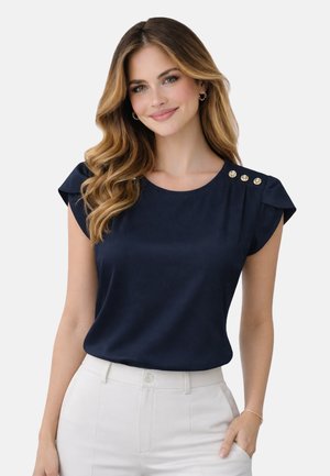 Frau mit langem, welligem Haar trägt ein marineblaues Oberteil mit drei goldenen Knöpfen auf der Schulter und weiße Hosen, lächelt leicht und hat die Hand in der Tasche.