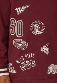 Bordeaux sweatshirt met witte geprinte graphics, waaronder logo's, een dinosaurusontwerp en verschillende patches. Witte ribbing aan de mouwen.