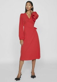 Červené šaty z ľahkej tkaniny, s dlhými puff rukávmi, hlbokým výstrihom do V a opaskom v pase. Hladká textúra; MIDI dĺžka.