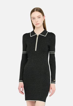 Robe noire côtelée avec un col blanc, devant à zip mi-long, et manches longues avec des accents blancs aux poignets, design ajusté.