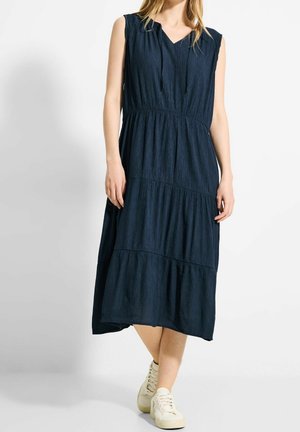 Kobieta w granatowej sukience midi bez rękawów z wielowarstwową spódnicą i dekoltem ze sznurkiem, w połączeniu z białymi tenisówkami, stoi na białym tle.