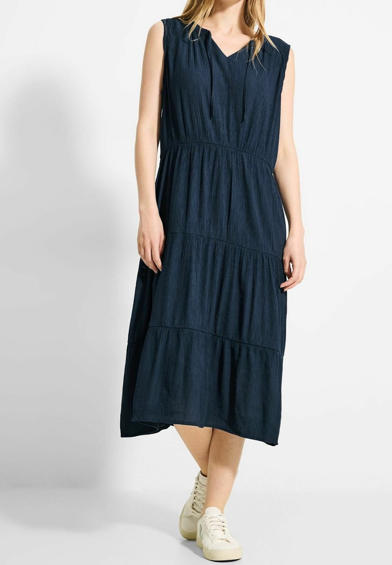 Femme portant une robe midi bleu marine sans manches avec jupe à volants et col à cordon, associée à des baskets blanches, debout sur fond blanc.