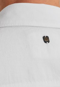 Camisa de cuello blanco con una tela texturizada, que presenta una pequeña etiqueta negra con "PME Legend" en dorado. Detalle de cuello clásico visible.