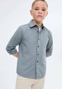 Poika, jolla on lyhyt hius, pukeutuneena harmaaseen nappipaitaan, jonka hihat ovat käärittynä, ja beigekat pantsit, seisoo kädet selän takana.