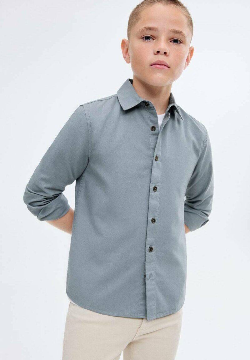 Poika, jolla on lyhyt hius, pukeutuneena harmaaseen nappipaitaan, jonka hihat ovat käärittynä, ja beigekat pantsit, seisoo kädet selän takana.