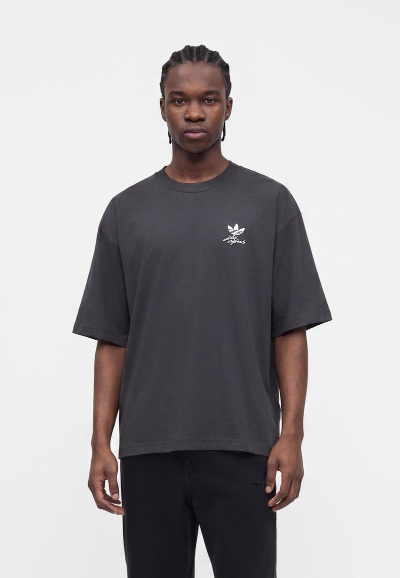 Junger Mann mit geflochtenem Haar, der ein lockeres dunkelgraues Adidas-T-Shirt und schwarze Hose vor einem schlichten hellen Hintergrund trägt.