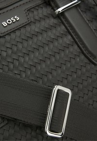 Zwarte geweven tas met een gestructureerd oppervlak, leren accenten, een stevige gesp en een merklabel. Compact ontwerp met een slanke uitstraling.