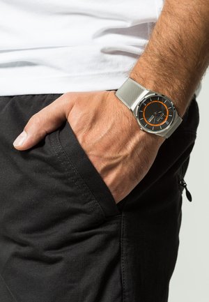 Männliche Hand mit silbernem Mesh-Armband-Uhr mit schwarzem und orangefarbenem Zifferblatt, die in der Hosentasche einer schwarzen Hose liegt.