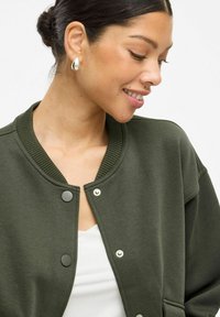 Tumši zaļš bomber jakas ar ribotu apkakli, spiedpogām un brīvu piegriezumu. Uzvilkts virs baltas blūzes, kurai ir gluda tekstūra.