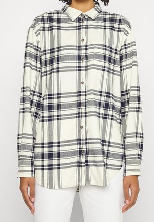 Koszula z długim rękawem zapinana na guziki w czarno-szarą kratę, noszona na białych spodniach, widok z przodu od szyi do bioder.