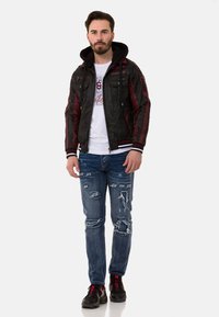 Schwarze und maroonfarbene Bomberjacke mit Kapuze, ausgestattet mit Reißverschlusstaschen, kombiniert mit ausgefransten blauen Jeans und schwarzen Sneakers mit roten Akzenten.