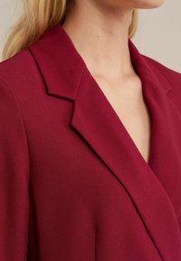 Blazer bordeaux avec une texture lisse, présentant un col à revers et un décolleté en V profond. Vue en gros plan du col et de la zone des épaules.