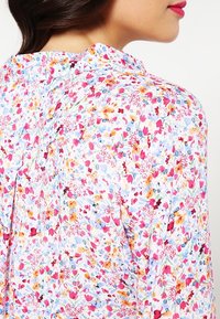 Blomsterbluse med en hvid baggrund og et farverigt mønster af rosa, blå og gyldne blomster; blød, let stof og et plisseret design.