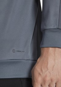 Maglietta sportiva grigia a maniche lunghe con tessuto testurizzato. Presenta il logo "AEROREADY" vicino all'orlo. Maniche lisce e design aderente.