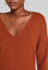 Maglione lavorato a maglia color ruggine con scollatura a V profonda, maniche lunghe e finitura strutturata, che mette in risalto una vestibilità morbida e rilassata.