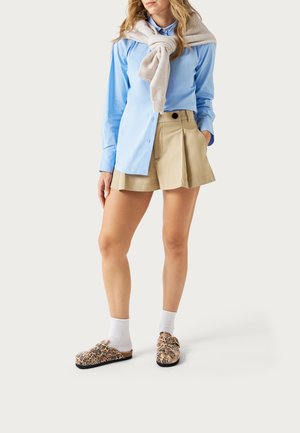 Mujer con camisa azul claro, pantalones cortos plisados beige, suéter beige sobre los hombros, calcetines blancos y zapatos sin cordones con estampado de leopardo.