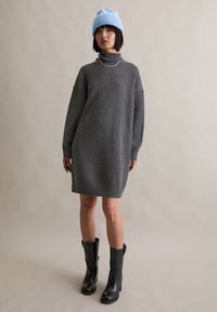 Grijze trui jurk met een hoge hals, geribde mouwen en zoom. Gecombineerd met zwarte leren laarzen tot aan de knie en een lichtblauwe gebreide beanie.