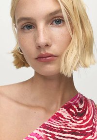 Capelli biondi di media lunghezza e occhi azzurri. Pelle liscia con lentiggini; indossa un indumento a spalla unica con un motivo ondulato rosa e bianco.