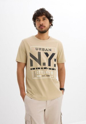 Homme aux cheveux bouclés portant un t-shirt graphique beige "Urban N.Y. Style" et un pantalon cargo clair, les mains dans les poches, sur fond blanc.