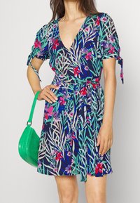 Květinové šaty s námořnickým pozadím a živými zelenými, růžovými a fialovými rostlinnými vzory, krátké rukávy a páskem v pase. Doplněné malou zelenou taškou.