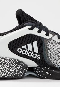 Czarne buty sportowe z siateczkową cholewką, białymi akcentami i nakrapianą czarno-białą podeszwą. Zawierają logo Adidas i fakturowe wzory.