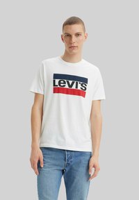 Joven con cabello corto que lleva una camiseta blanca de Levi's con rayas azul marino y rojas, y jeans azules, contra un fondo neutro.