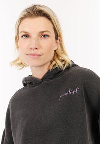 Donkergrijze hoodie met een zachte textuur, voorzien van een kangoeroezak en een roze geborduurd "protest" logo op de borst.