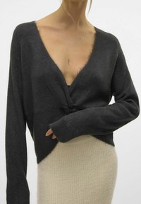 Ciemnoszary krótki sweter z głębokim dekoltem w serek, długimi rękawami i zawiązanym na przodzie detalem, zestawiony z jasnobeżową dzianinową spódnicą.
