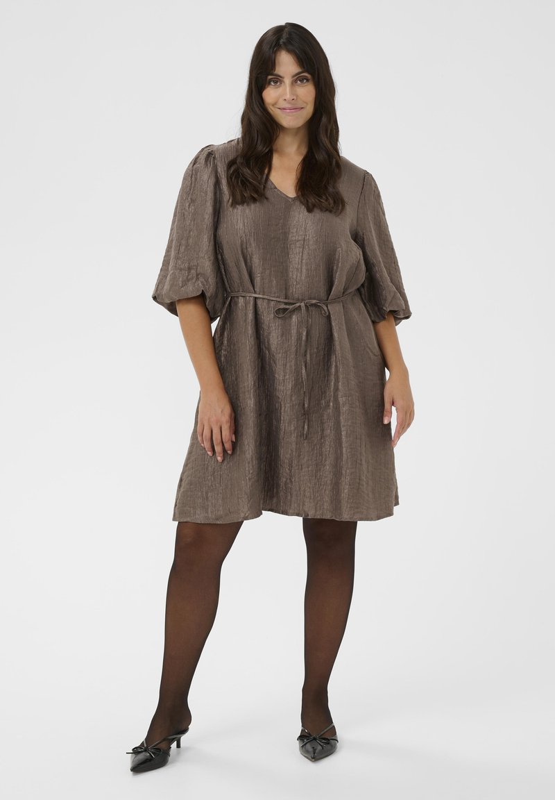 Frau stehend in einem strukturierten braunen knielangen Kleid mit Puffärmeln, schmalem Gürtel, transparenten schwarzen Strumpfhosen und schwarzen spitzen Absätzen mit Schleifen.
