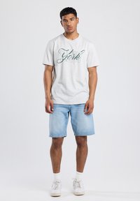 Grijze katoenen t-shirt met groene "New York" graphic, gecombineerd met lichtblauwe denim shorts en witte sneakers, met witte sokken.