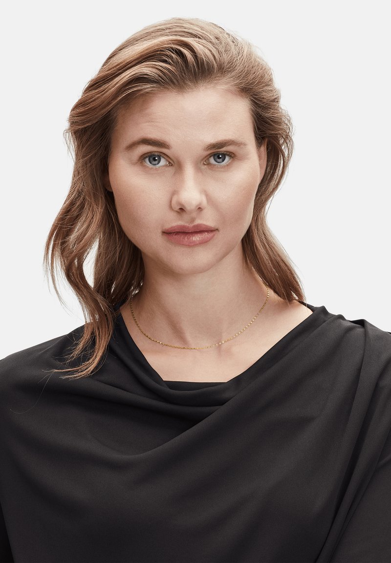 Collier en or avec de délicates petites perles rondes, porté sur un haut en tissu drapé noir. L'arrière-plan est blanc uni.