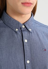 Camisa de botones azul oscuro con una tela texturizada, con cuello amplio y botones blancos. Pequeño logo en el lado izquierdo.