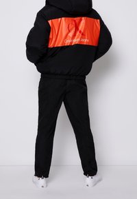 Svart puffjacka med ett orange horisontellt streck som har "Calvin Klein Jeans"-logotypen. Kombination med svarta byxor och vita sneakers.