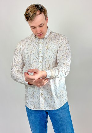 Jeune homme portant une chemise blanche à fleurs boutonnée et un jean bleu, ajustant sa manche gauche sur un fond clair uni.