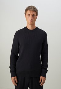 Man standing against plain background wearing black ribbed long-sleeve sweater and black pants, looking straight ahead with neutral expression.