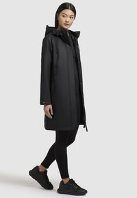 Zwarte waterdichte jas met capuchon, gekenmerkt door een rechte snit, drukknoopsluiting en ritszakken aan de zijkanten. Zwarte leggings en sportschoenen maken de look compleet.