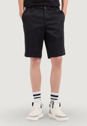 Persona che indossa pantaloncini neri con le mani in tasca, scarpe da ginnastica bianche alte e calzini bianchi con strisce nere.