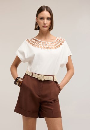 Vrouw met kort bruin haar die een witte top met ronde uitsnede draagt, bruine shorts met hoge taille, een geweven riem en armbanden, staand met handen in de zakken.