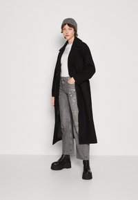 Sort lang frakke over en hvid top, parret med grå denim cargo bukser med malingsplætter. Sorte platform støvler med en tekstureret beanie.