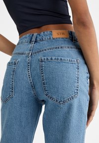Jeans de mezclilla azul claro con cintura alta, pierna recta y dos bolsillos traseros. Presenta una parche de cuero marrón con "STR" en la cinturilla.