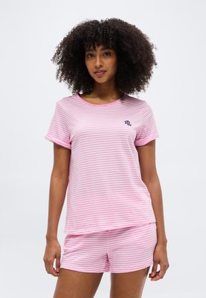 Kvinde med krøllet hår iført lyserødt og hvidt stribet kortærmet top og matchende shorts, stående mod en ensfarvet lys baggrund.