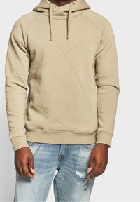 Beige hættetrøje med skrå søm, lavet af en bomuldsblanding, med ribbede cuffs og snøre i hætten, parret med slidte blå jeans.