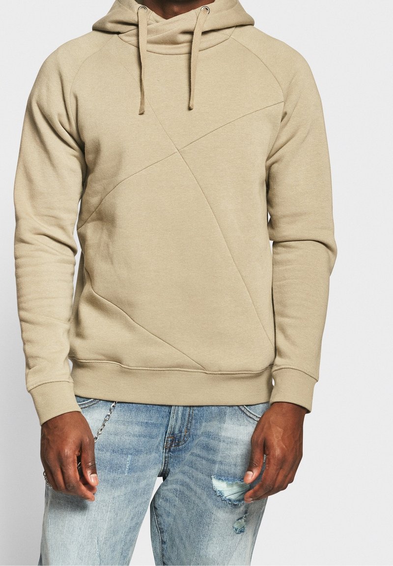 Beige hættetrøje med skrå søm, lavet af en bomuldsblanding, med ribbede cuffs og snøre i hætten, parret med slidte blå jeans.