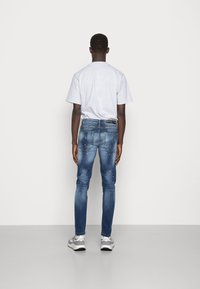 Jeans azules de hombre con un corte ajustado, detalles desgastados y un lavado medio. Combinados con una camiseta de manga corta gris y zapatillas grises.