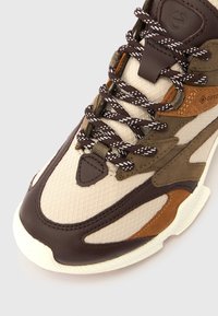 Zapatilla beige y marrón con acabado de malla, acentos de cuero, lengüeta acolchada, suela texturizada y cordones contrastantes en marrón y blanco.