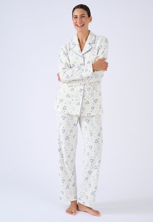 Mujer sonriendo con los brazos cruzados, usando pijama blanca de manga larga abotonada y pantalones con estampado de corazones azules y grises.