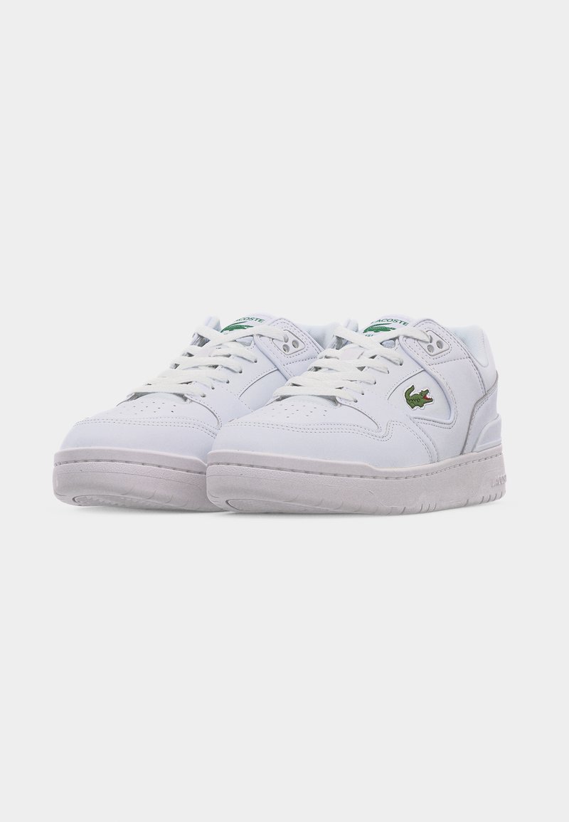 Witte lage Lacoste sneakers met veters, geperforeerd teengebied en groen krokodillenlogo aan de zijkant, weergegeven tegen een effen achtergrond.